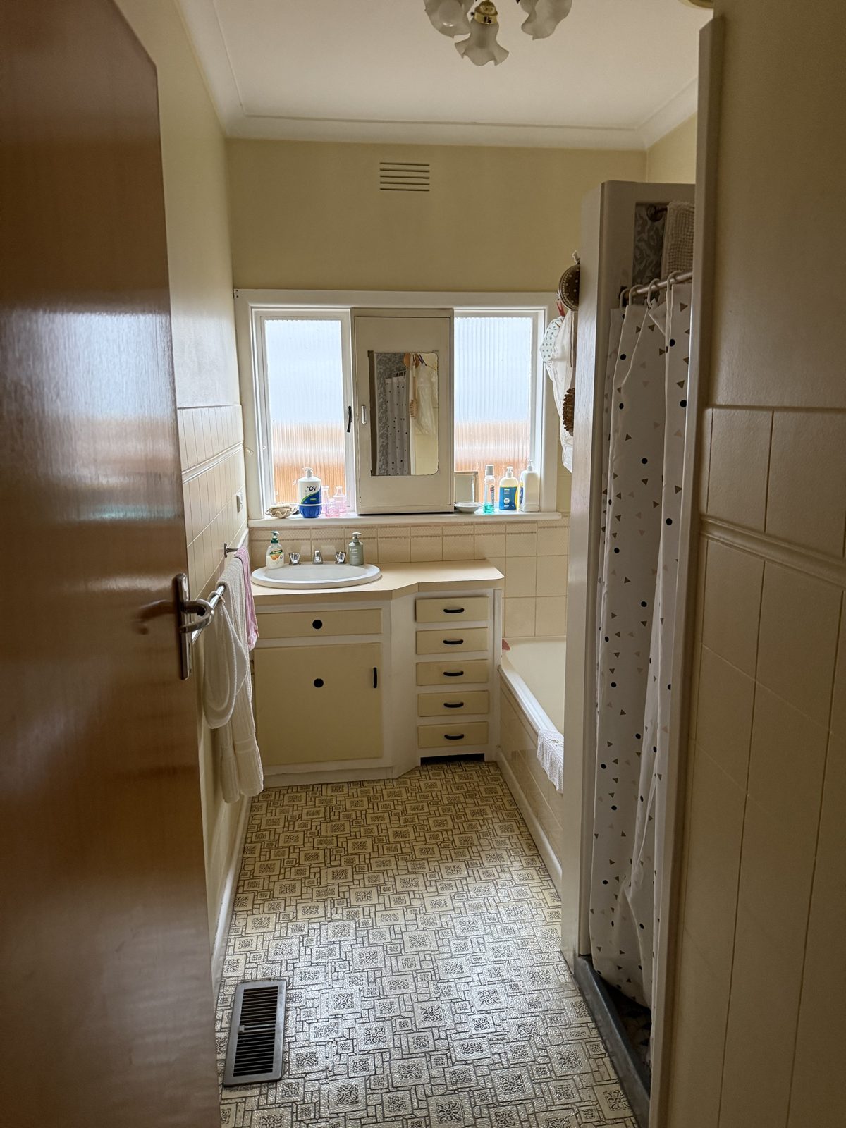 The original 1960s bathroom in Altona North before renovation: cream tiles, patterned vinyl floor, bath with a curtained shower